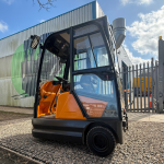 STILL R6-06 tow tractor in black and orange, featuring a fully enclosed cabin with glass doors. Ideal for indoor and outdoor towing in warehouses, factories, and logistics operations.